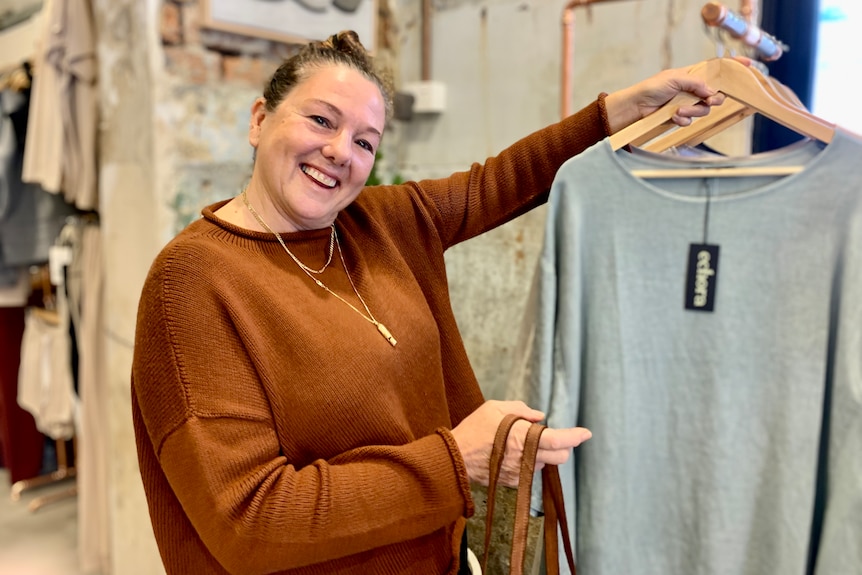 Shop owner Donna Cooper smiling holding a coat hanger with grey top 