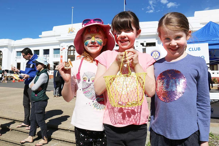 three little girls with chocolate eggs and painted faces stand outside Parliament House.