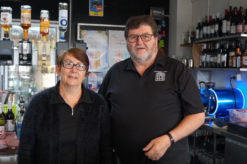A man and a woman in black clothes and glasses behind a pub bar.