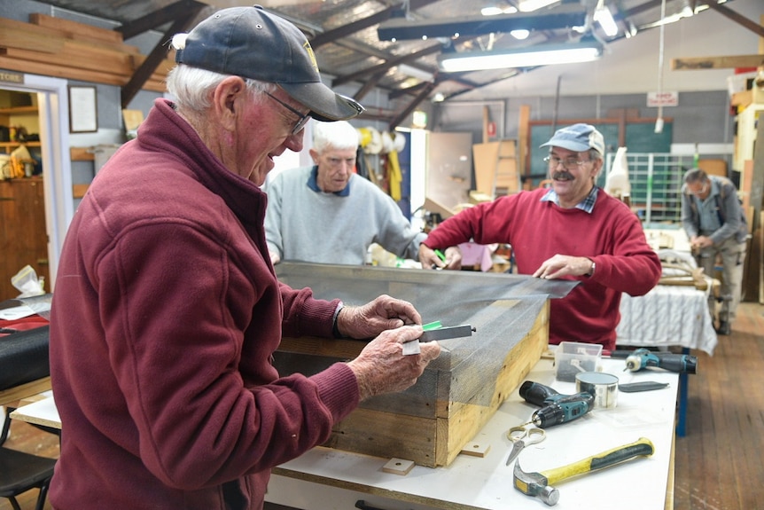 Three men hammering mesh onto a square wooden frame on a table in a workshop.