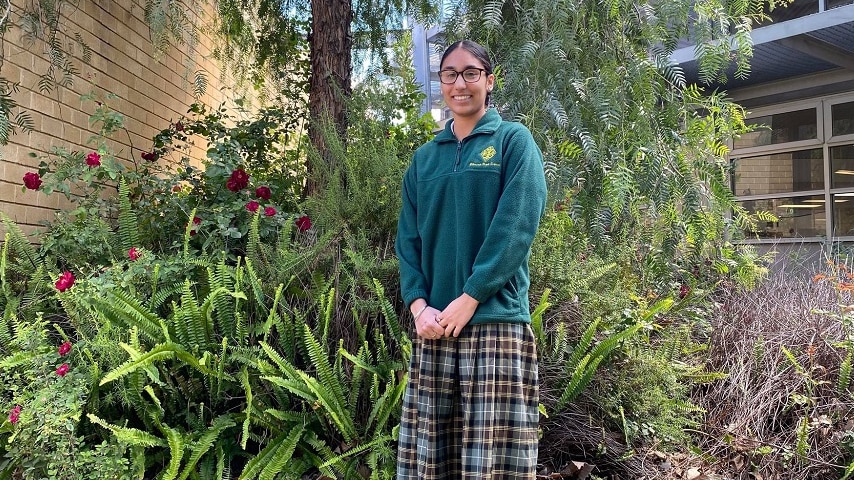 A woman is standing and smiling in front of a tree and bush. There are maroon flowers on the bush.