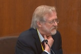 Martin Tobin sits in a wooden witness box grabbing his throat as he looks to the jury box. 