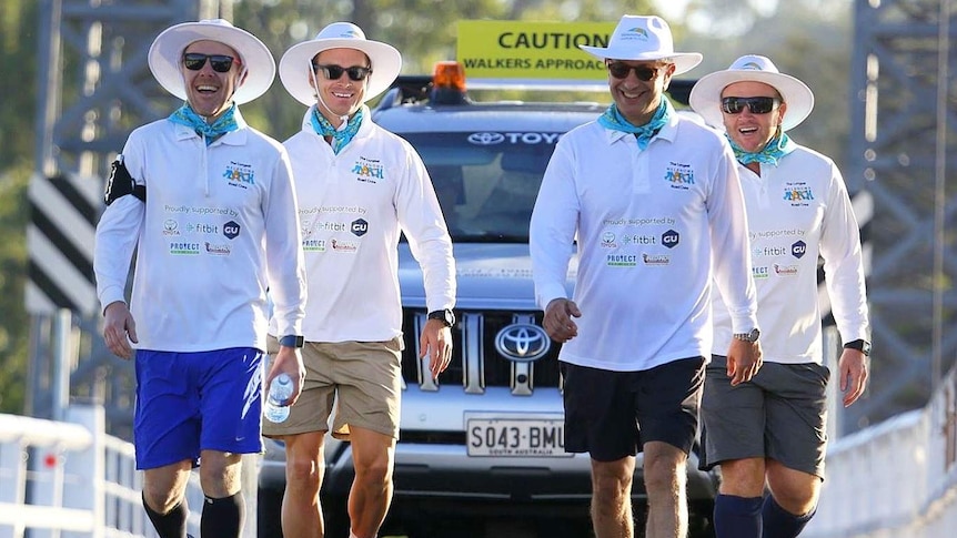 Four men walking across a bridge with a support vehicle behind them.