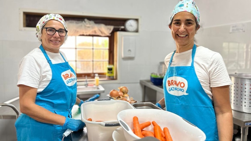 Two women wearing colourful hair nets and bright blue aprons smiling while working in a kitchen.