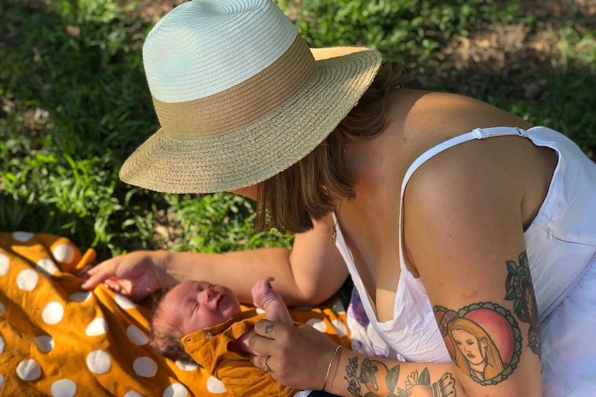 A woman bends down and holds the arm of a baby. She is wearing a white dress and has a hat on. The baby wears orange.