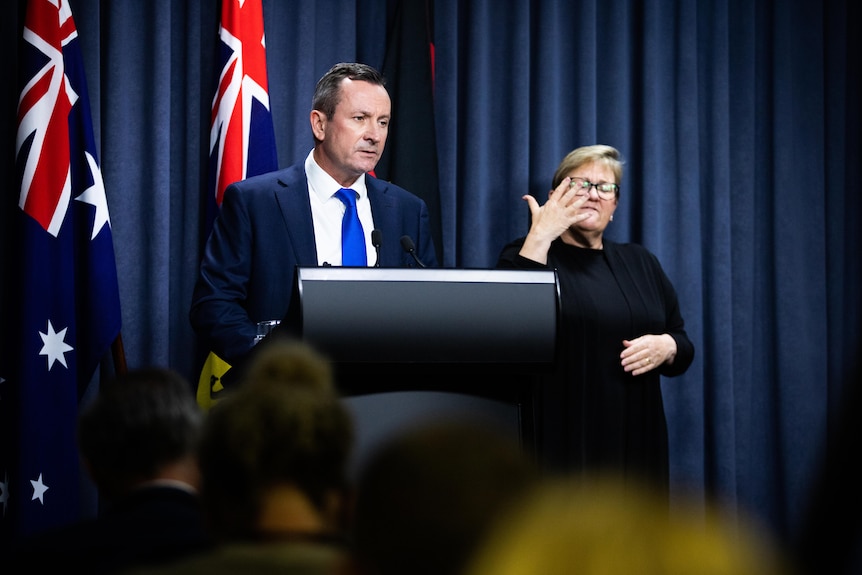Man speaks at lectern in front of Australian, Western Australian, and Aboriginal flags, with an Auslan interpreter at his side