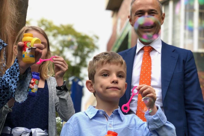 Little boy stands in foreground blowing bubbles while girl blows bubbles in background and man watches on