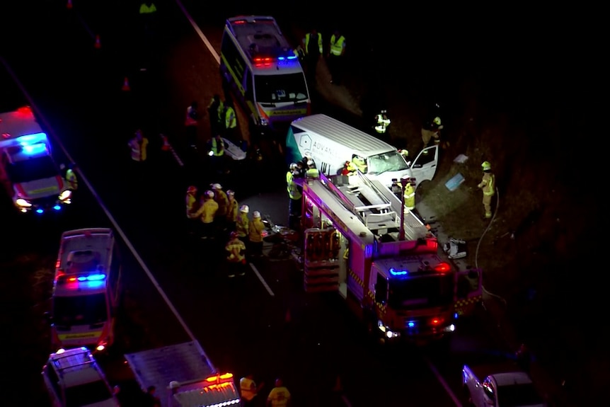 Emergency services vehicles with flashing lights surround a white van with workers trying to free the driver.