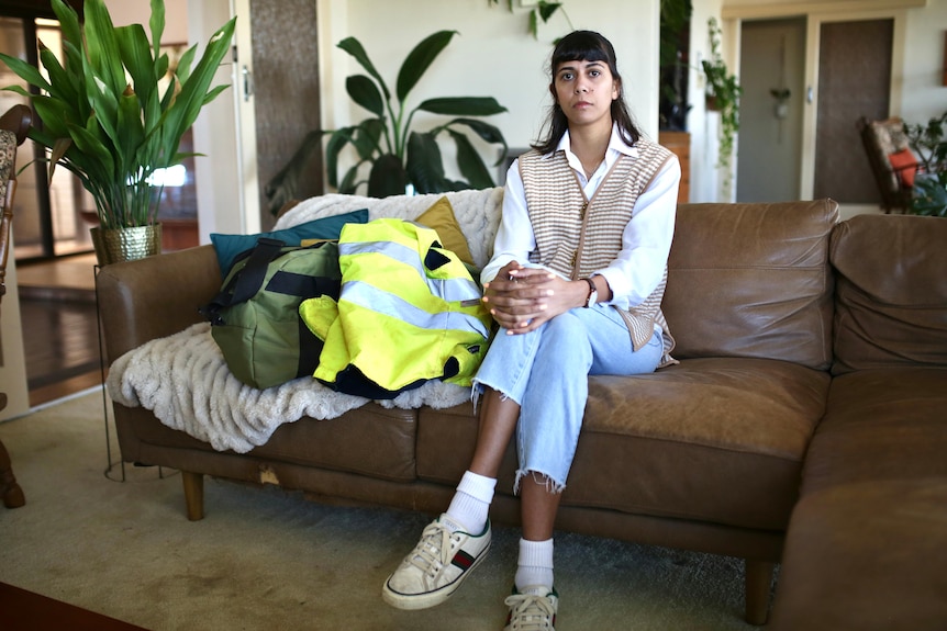 A wide shot of a woman sitting on a couch indoors with her legs crossed and a bag and hi-vis jacket next to her.