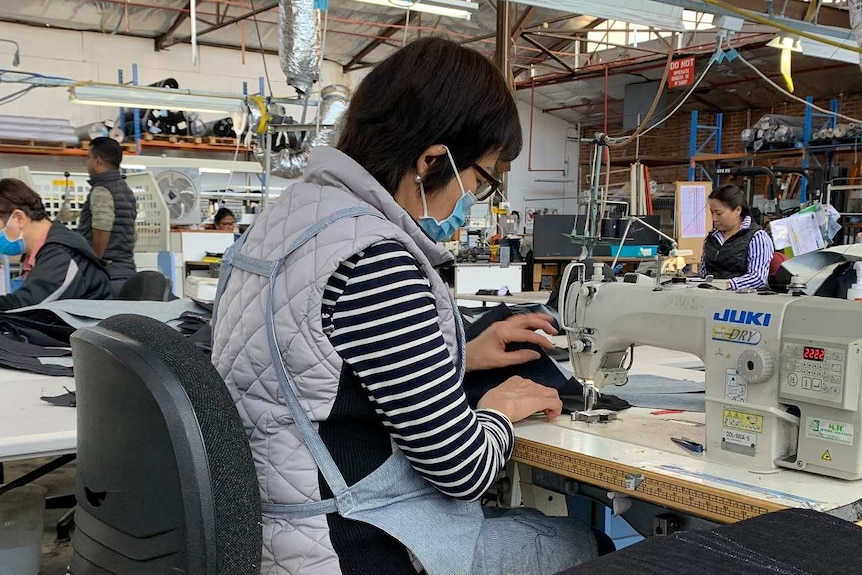 A woman sits over a sewing machine making jeans in a factory surrounded by other workers bent over sewing machines.