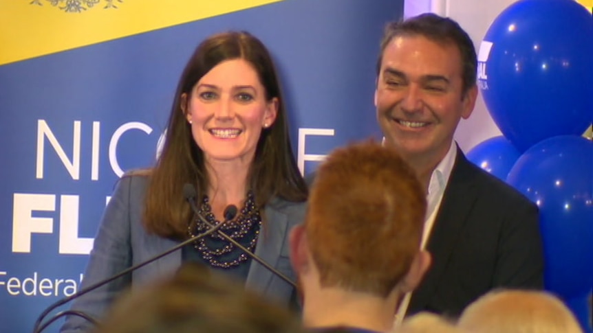 A woman and a man in front of a sign saying Nicolle Flint and blue balloons