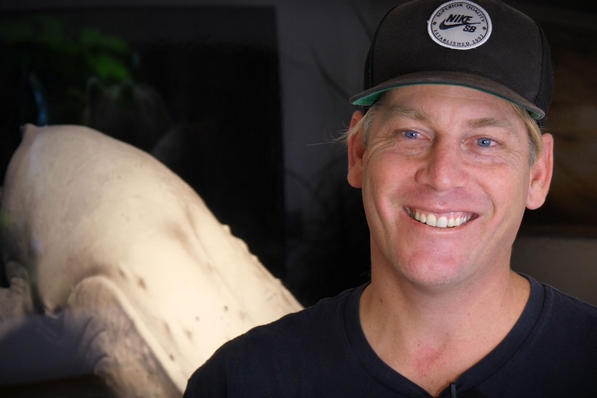 A man in a black cap and shirt is smiling in front of an image of a white whale on a black background