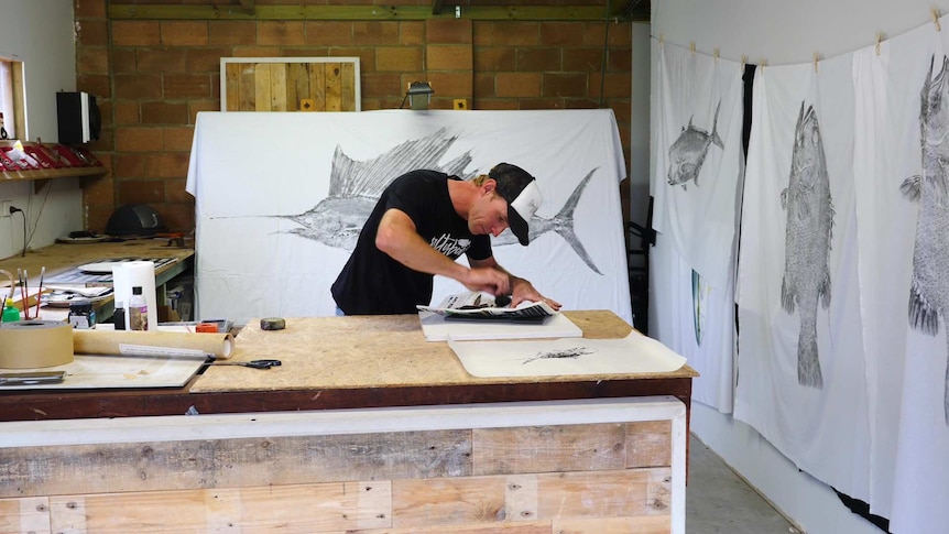 A man in a black shirt and cap bends over a bench surrounded by fish artwork in a room.