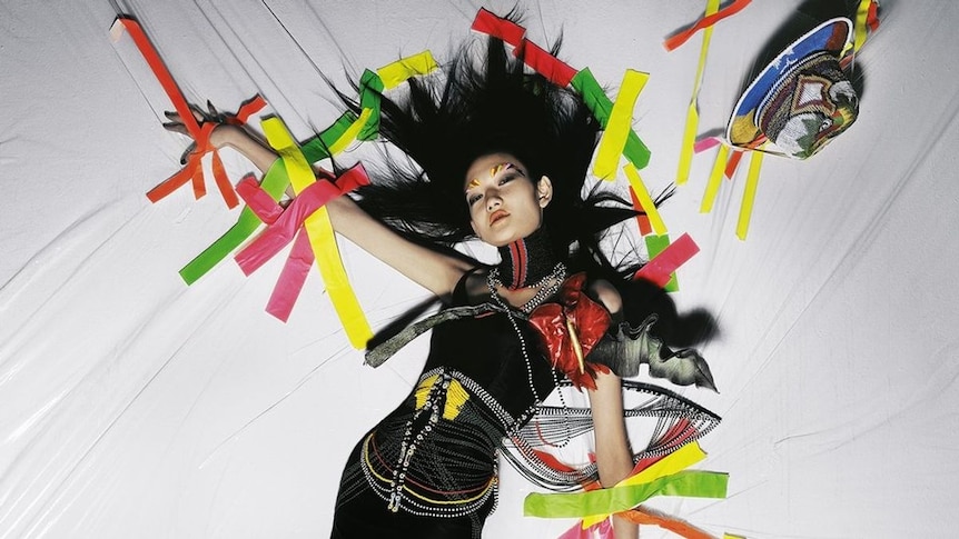 A fashion model is held to a white sheet with multi coloured sticky tape