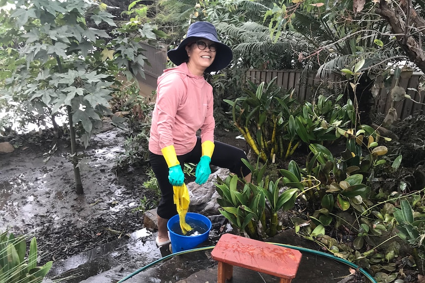 A woman wearing rubber dishwashing gloves dips a yellow dishcloth into a bucket of murky water.