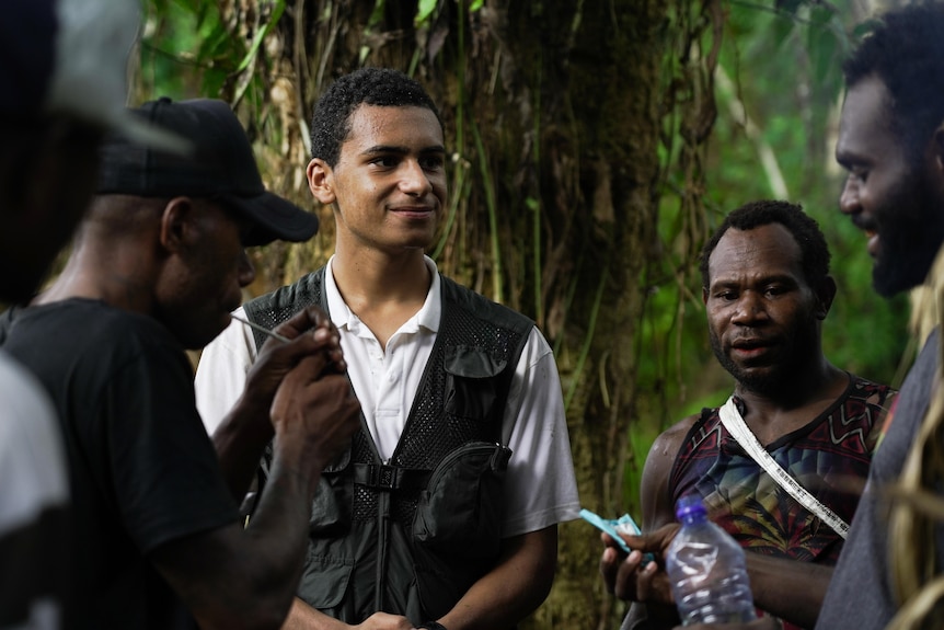 A group of young men stand together in front of a tree in dense jungle