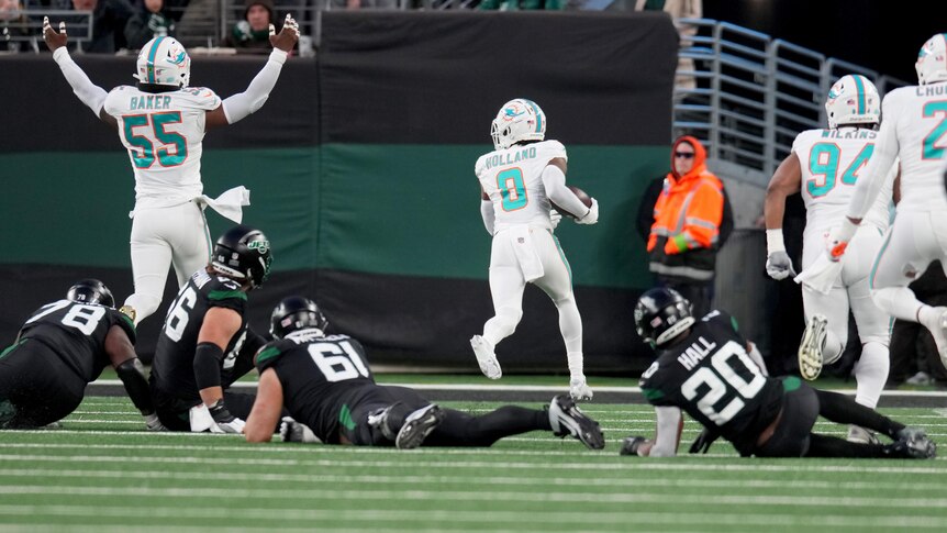 A Miami NFL player runs the ball into the end zone, as his teammate raises his arms, and opposition players lie on the ground.