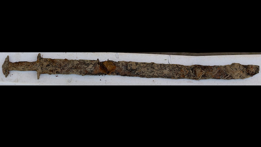 A sword covered in what looks like mud or thick rust is lying on a white plank, on a black background