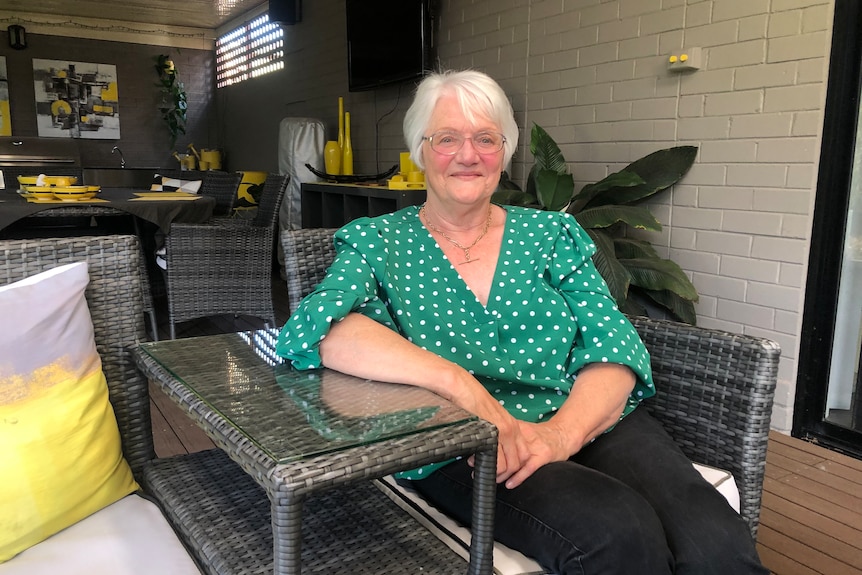 Marilyn Baker, with short, white hair, bright green blouse and black pants, sits in an outdoor chair smiling with mouth closed.
