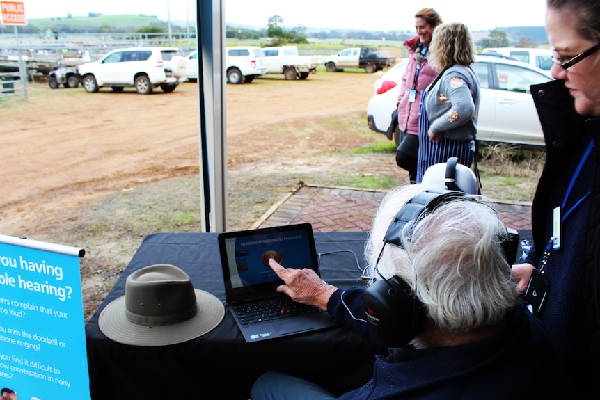 A man with white hair wearing headphones and pressing an iPad resting on a table, woman over shoulda