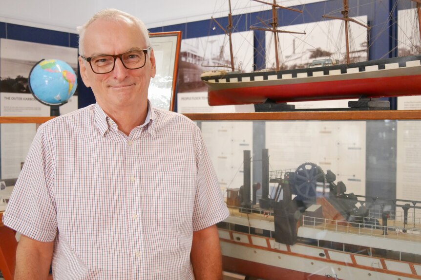A man in a checkered shirt and glasses, smiles in front of a model of a boat