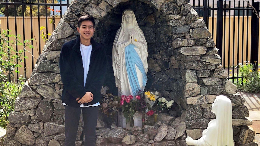 Hieu standing in front of a rock grotto with a statue of the Virgin Mary inside.