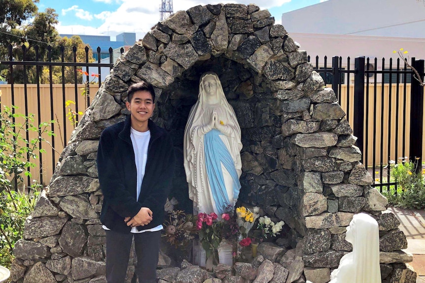 Hieu standing in front of a rock grotto with a statue of the Virgin Mary inside.