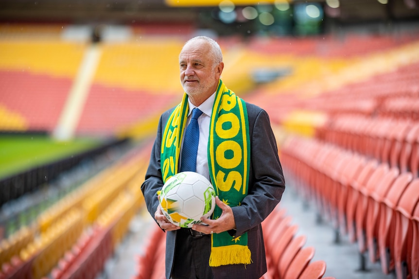 A grey-haired man wearing a Socceroos scarf and holding a soccer ball stands in an empty stadium, looking off towards the field