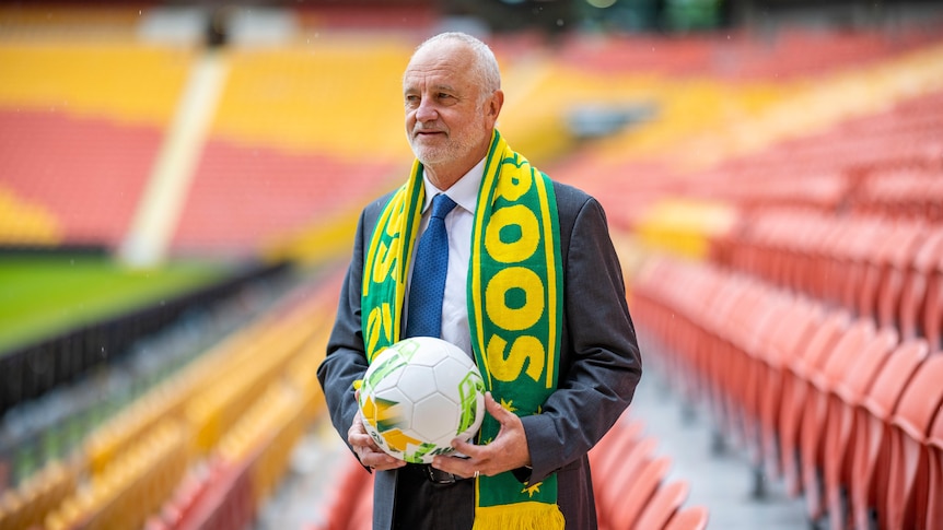 Graham Arnold stands in an empty stadium, wearing his green and yellow Socceroos scarf and carrying a branded soccer ball.
