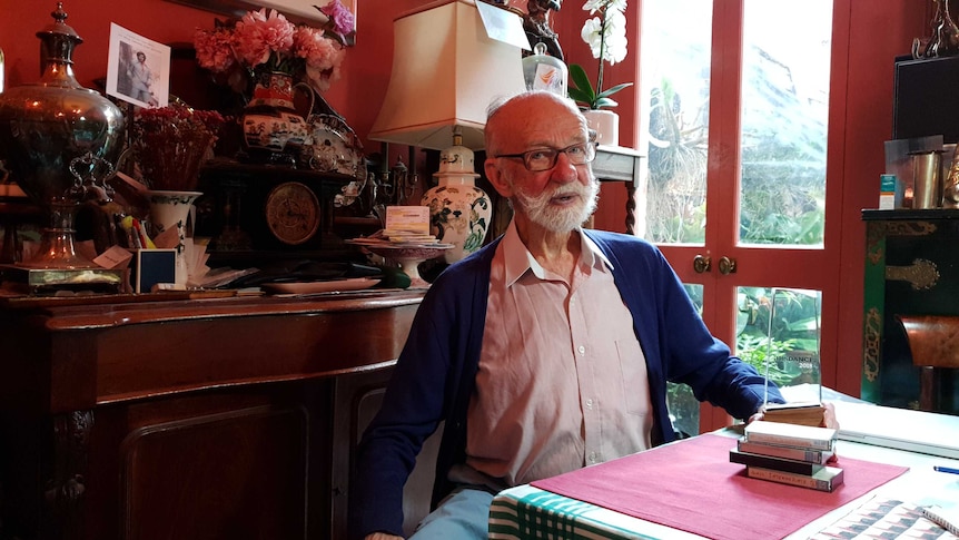 Athol Willoughby sits at a table in a red living room. An Australian Dance Award and a pile of cassette tapes are on the table.