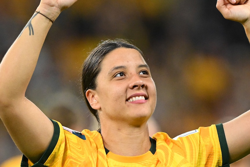 Sam Kerr raises her hands above her head as she celebrates and smiles.