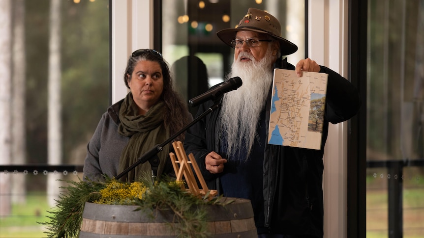 A woman and a man stand next to each other in behind a wooden barrel. The man wears a hat and has a beard, and holds a map. 