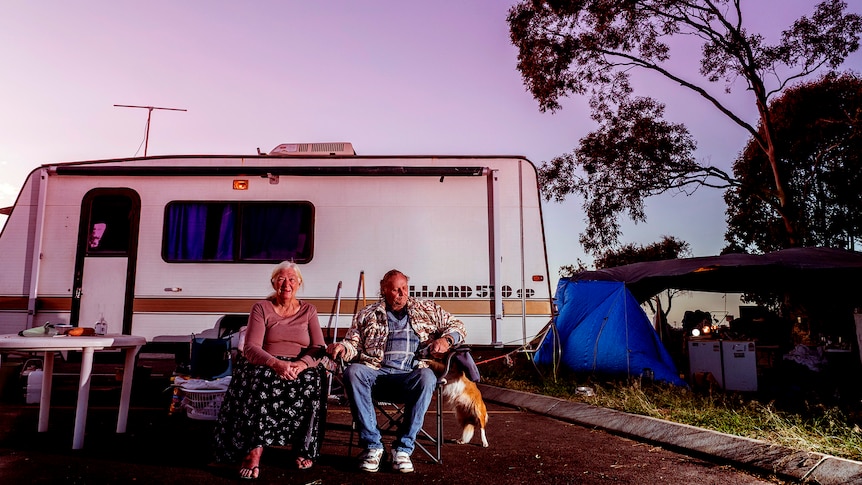 A woman and man sit outside a caravan with a dog.