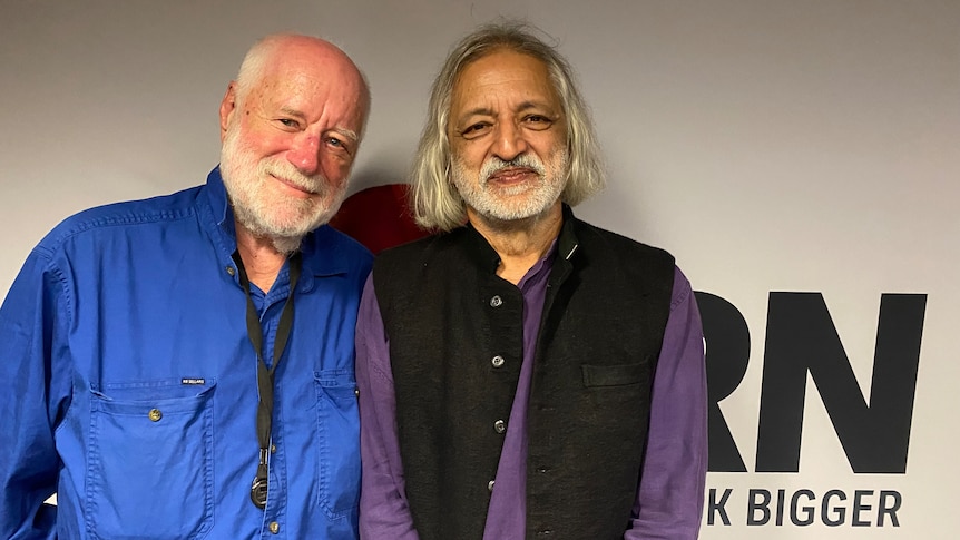 An older Indian Man stands in front of the RN logo with an old white man with a beard