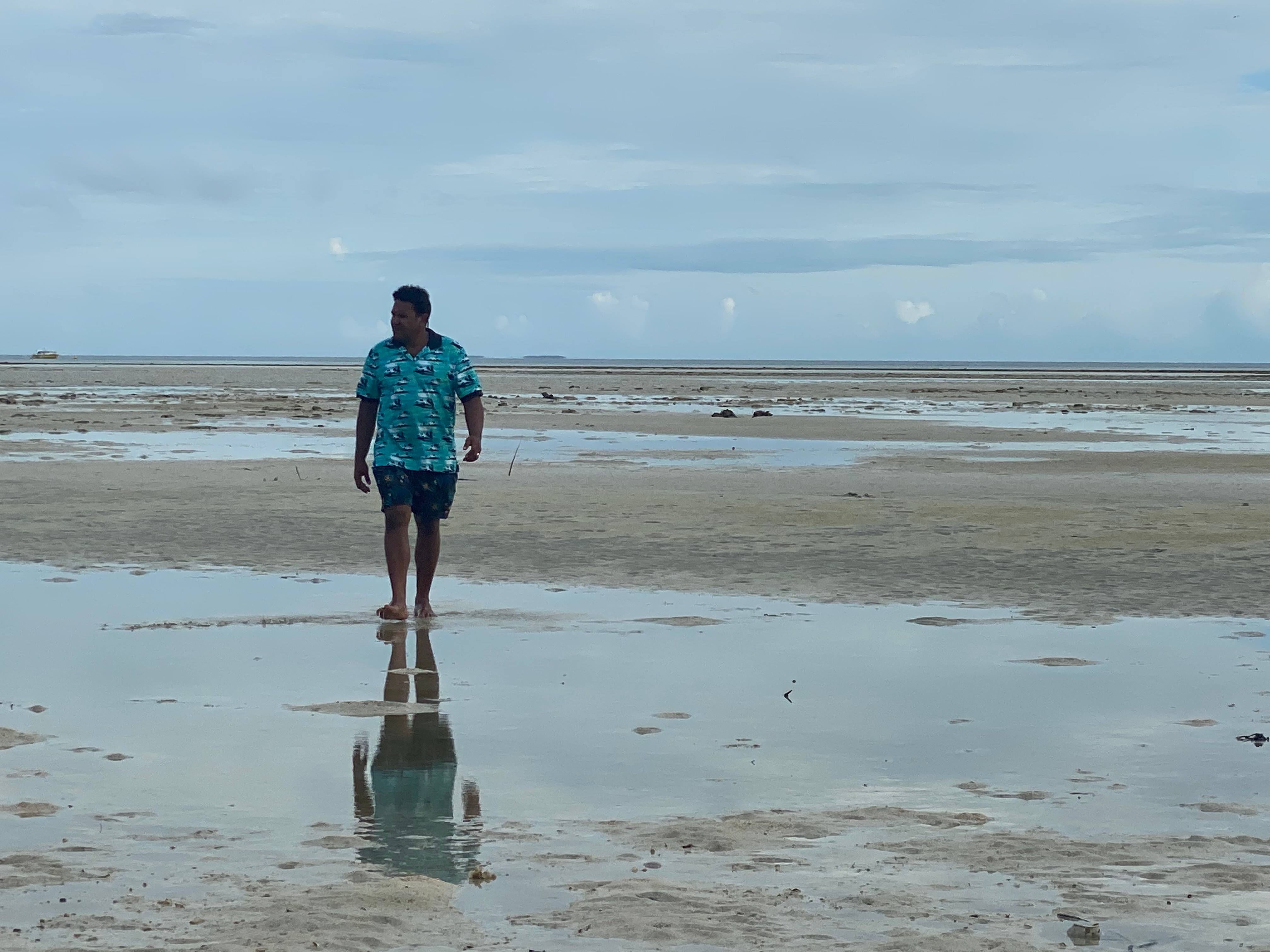 Man in blue shirt and shorts walks along the beach.