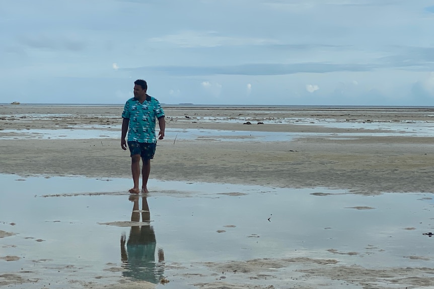 Man in blue shirt and shorts walks along the beach.