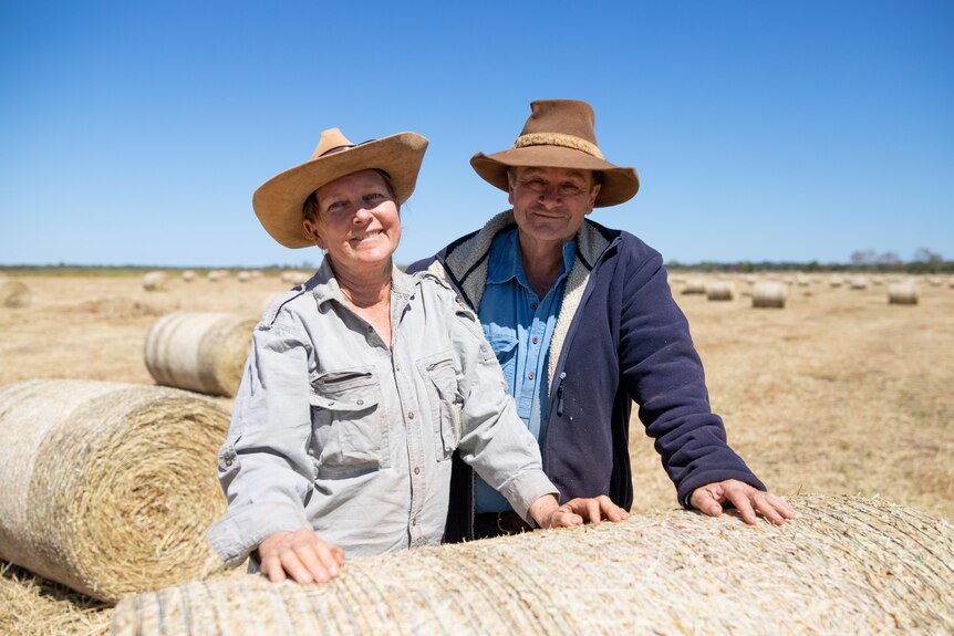 A middle-aged man and woman lean on hay bale in a paddock and smile at the camera, wearing broad-brimmed hats and work shirts 