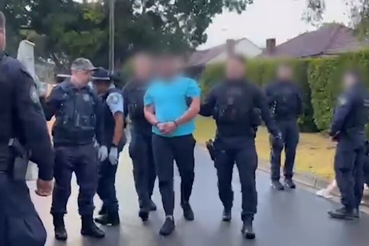 A man with his face blurred is flanked by several police officers as he's led to a police van