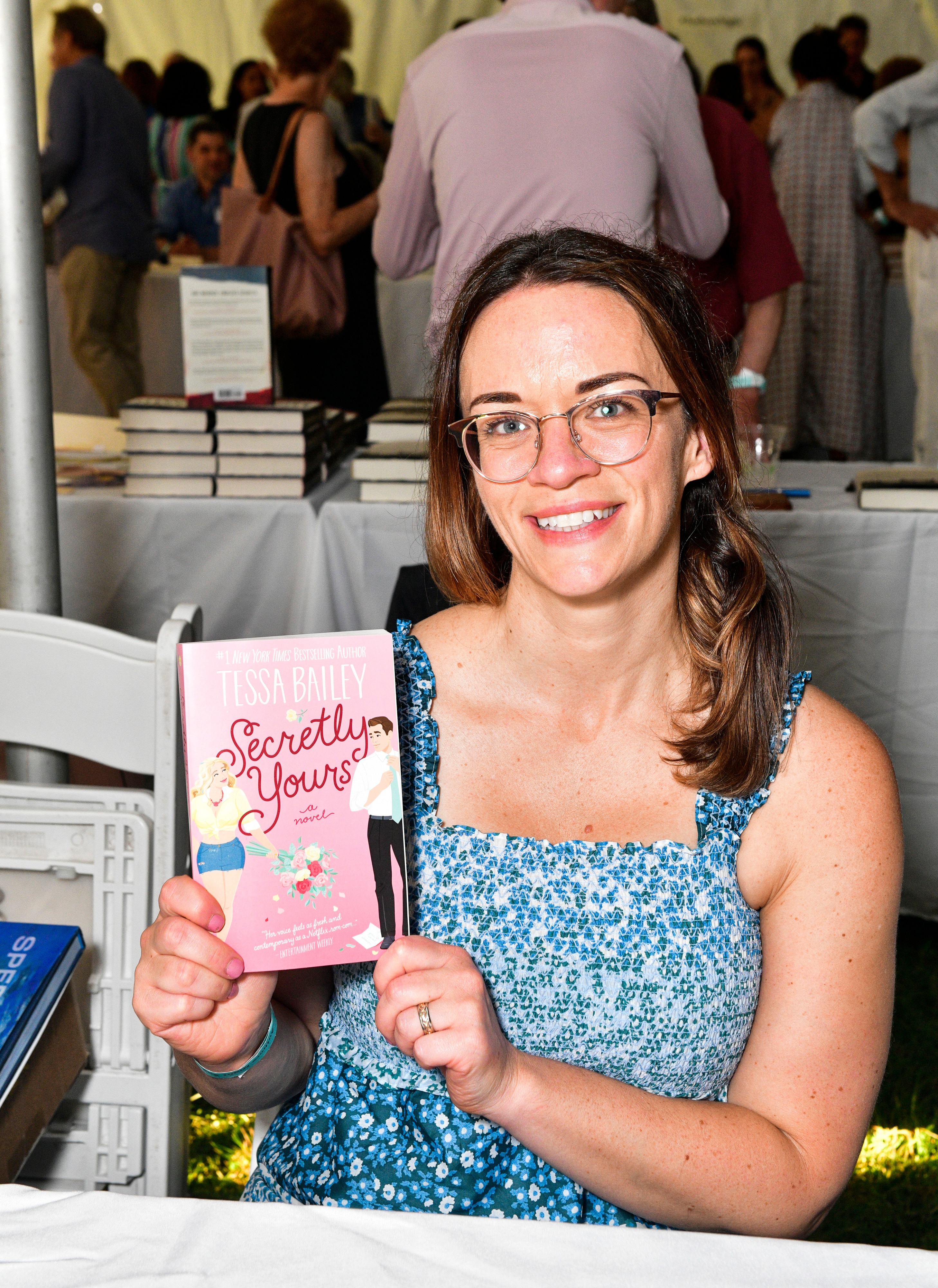 Tessa, who has brown hair and wears a blue dress and glasses, smiles as she holds up a pink romance novel.
