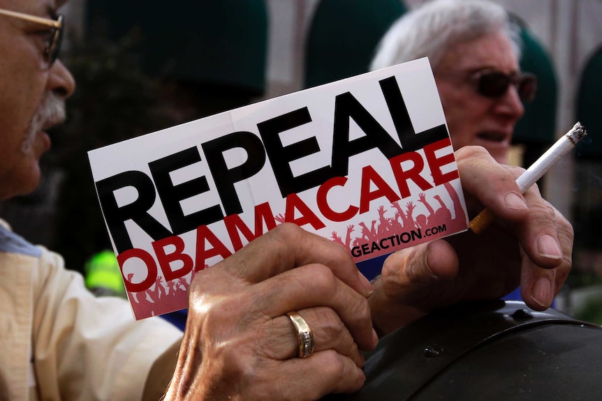 A man's hand holds a small sign reading "Repeal Obamacare" at a demonstration in Indiana 2013.