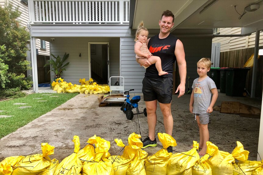 A man holds a toddler with his young son standing next to him, with sandbags around them.