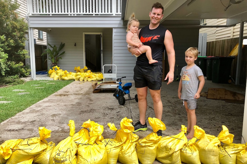 A man holds a toddler with his young son standing next to him, with sandbags around them.