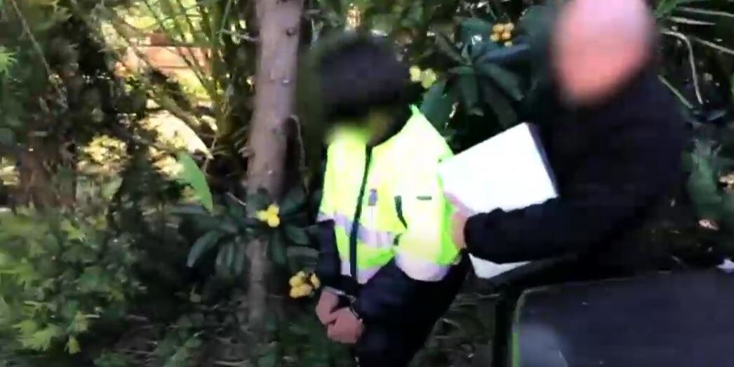Police officer walks with man in handcuffs, both faces blurred.