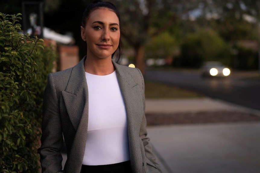 A woman wearing a white top and a checked blazer stands next to a pruned hedge.