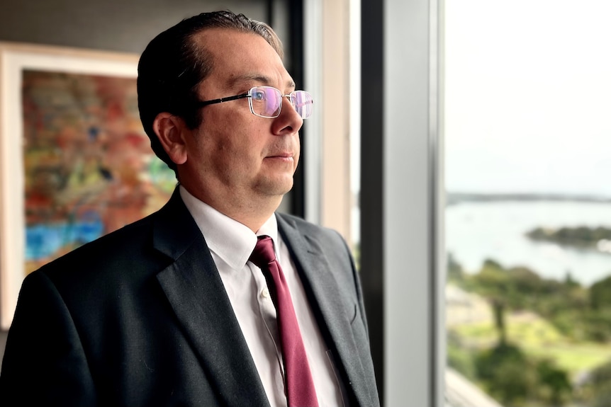 A man with brown hair and glasses wearing a suit with a red tie sits at a board table, looks out a window