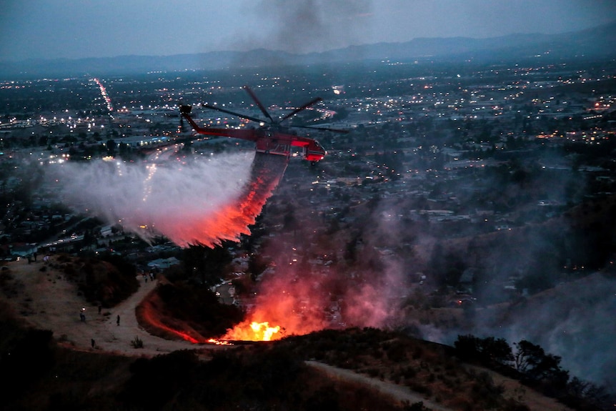 A large helicopter drops water above homes in California, the sky is dark and the glow of the fire reflects on the helicopter.