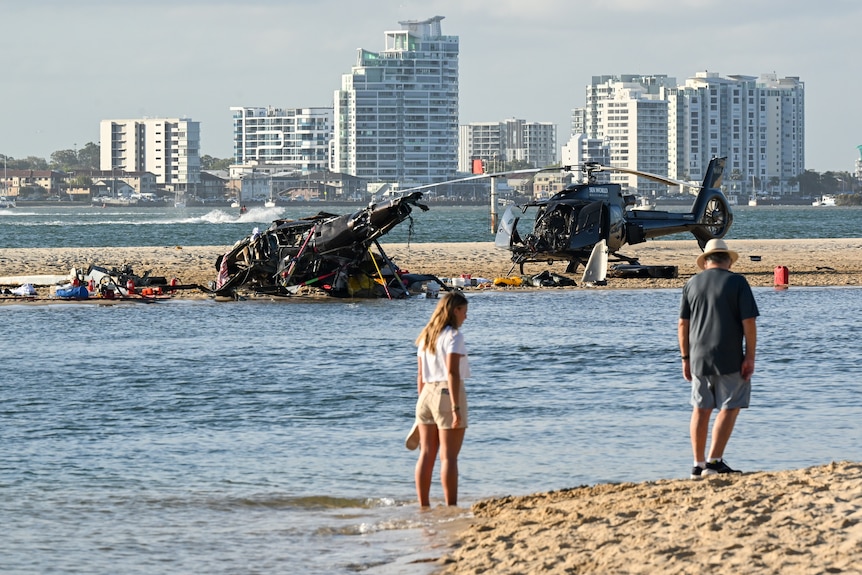 Two people stand on a beach near the wreckage of two helicopters.