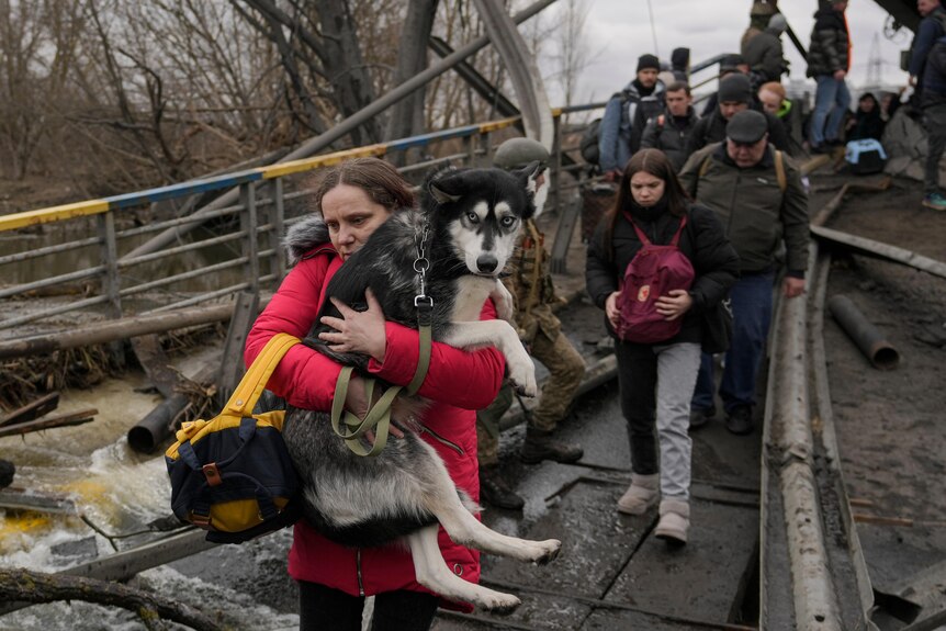 a woman holds a medium to large sized dog in her arms as she follows a path under a bridge with debris around the path
