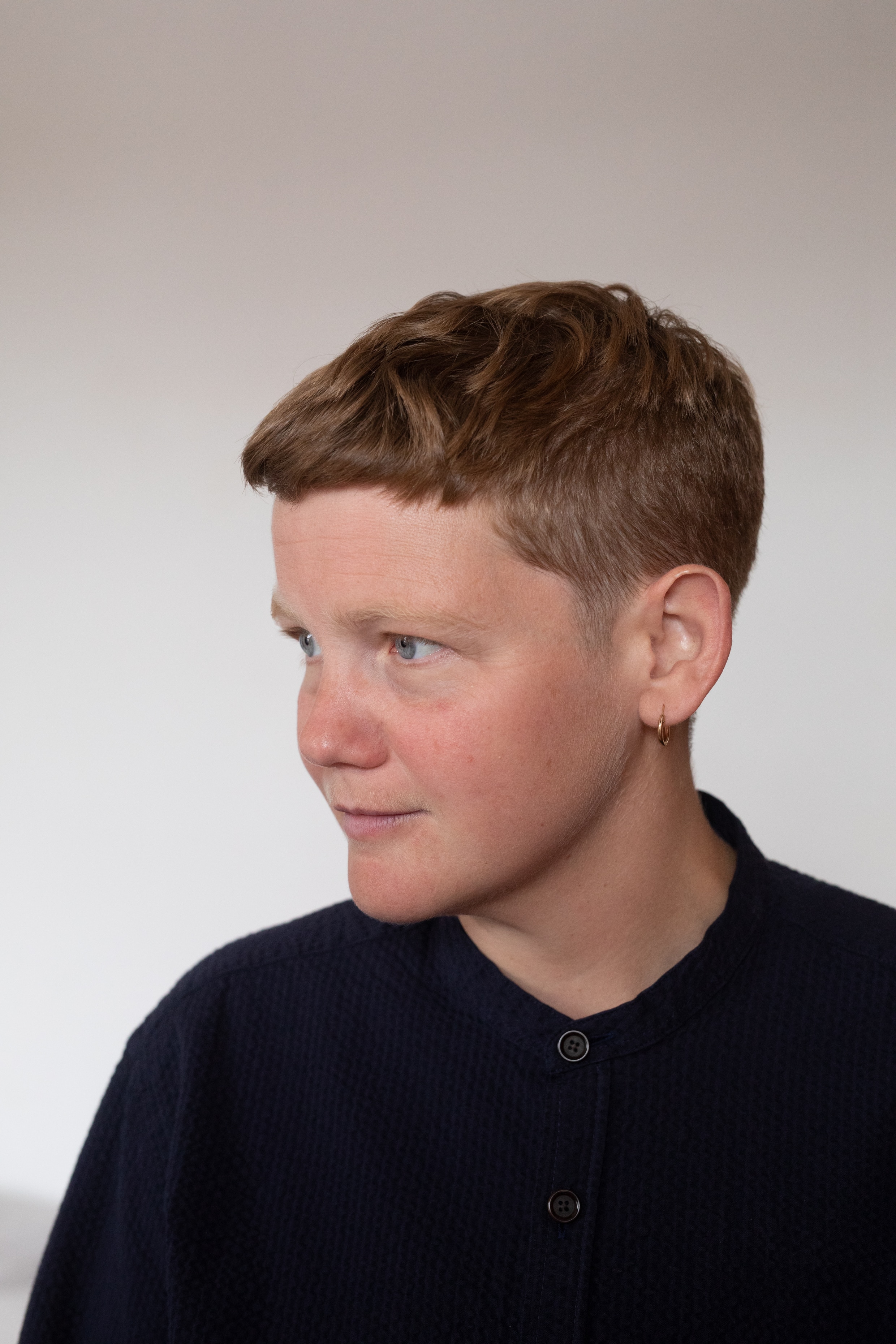 A white non-binary person with short sandy hair and blue eyes wears a high-necked navy shirt and looks off-camera.