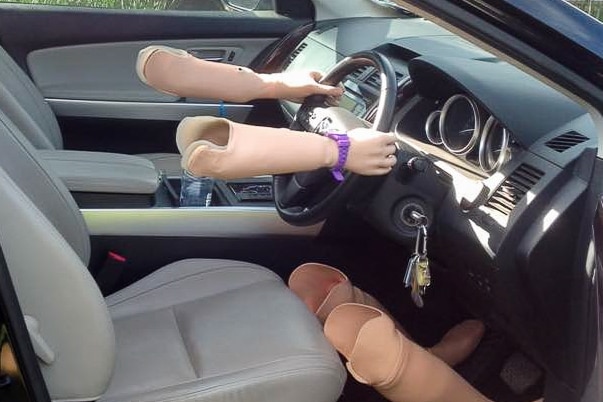 Two prosthetic arms on a steering wheeel, with two prosthetic legs standing up beside the driver's seat.